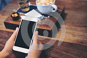 A woman`s hands holding white mobile phone with blank black screen with coffee cup , tea and snack on wooden table in vintage caf