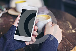 Woman`s hands holding white mobile phone with blank black screen and a coffee cup on wooden table in vintage cafe