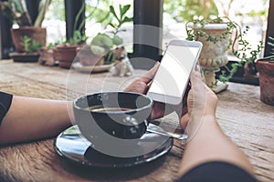 Woman`s hands holding white mobile phone with blank desktop screen and coffee cup on table in cafe