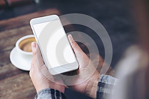 A woman`s hands holding white mobile phone with blank desktop screen with coffee cup on wooden table in cafe