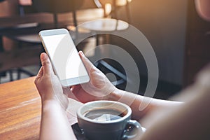 A woman`s hands holding white mobile phone with blank desktop screen with coffee cup on wooden table in cafe