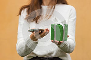 Woman& x27;s hands holds smartphone and green gift box with bow. Female in white sweater using mobile app and shopping
