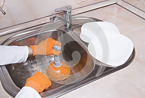WomanÃ¢â¬â¢s hands with orange gloves cleaning sewer and water at kitchen faucet over metal sink. Female hand with plunger. Home