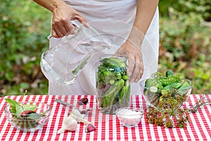 Woman`s hands pour water from decanter into jar of cucumbers for preservation