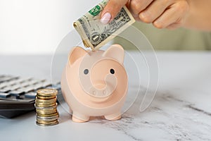 a woman\'s hands put dollar paper money in a piggy bank, next to a calculator on the table. economy, crisis and