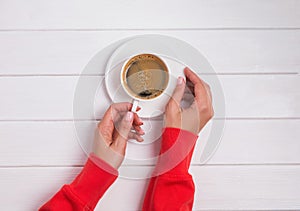 Woman`s hands in red clothing holding a cup of coffee on the white wooden table