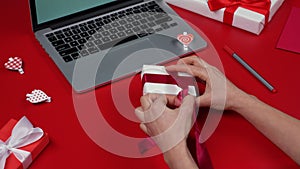 Woman's hands unpack the gift box, undoing bow. Top view of red table with laptop, gifts, hearts and valentine. Romantic