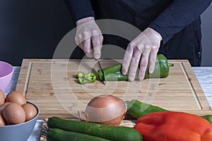 Woman`s hands using a knife to cut green pepper on a wooden board. Blurred, gray bowl with eggs, red and green peppers, onion and