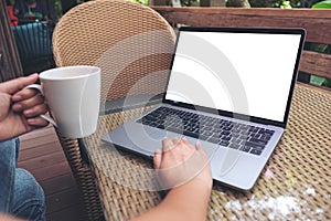 A woman`s hands using laptop with blank white desktop screen while drinking hot coffee on wooden table in cafe