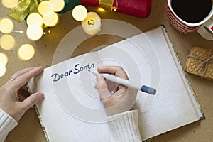 Woman`s hands in white sweater writing letter to Santa in notebook, selective focus, bokeh lights, atmospheric photo. Christmas