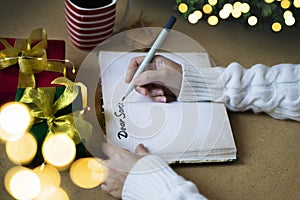 Woman`s hands in white sweater writing letter to Santa in notebook, selective focus, bokeh lights, atmospheric photo. Christmas