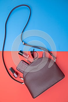 Woman\'s leather handbag in crimson color and accessories on red and blue background. Top view