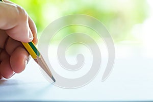 Woman left Hand holding multicoloured pencil Yellow & Green colour, writing letter on white paper, Bokeh green garden