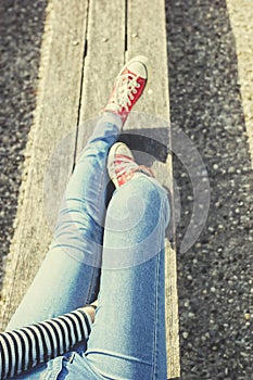 Woman`s legs in a blue jeans and red canvas sneakers sitting on a bench