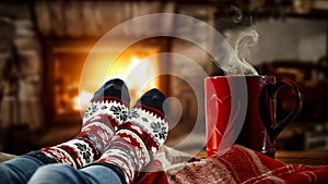 Woman`s legs with christmas socks and a red mug of coffee or tea and home interior with fireplace and dark wall background.