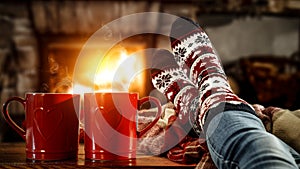 Woman`s legs with christmas socks and red mugs of coffee or tea and home interior with fireplace and dark wall background.