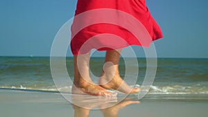 Woman`s legs close up. The ocean on a background. The woman in a red dress walks on the beach.