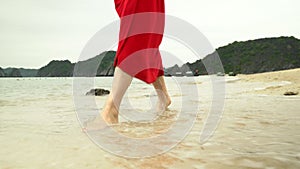 Woman`s legs close up. The ocean on a background. The woman in a red dress walks on the beach. Closeup of beautiful girl