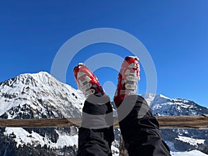 Woman& x27;s legs in red ski boots resting on wooden fence after a ski tour on sunny day, snowy mountains in the