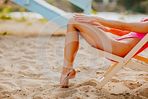 Woman& x27;s long slim legs in pink bikini sitting at the chair on the sand beach