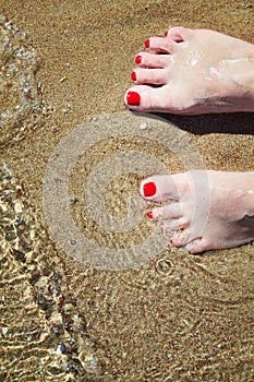 Woman`s pedicured feet with red nail polish on toes in the sand in water.