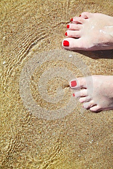 Woman`s pedicured feet with red nail polish on toes in the sand in water.