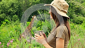 a woman in a safari hat drinks water near a giraffe in the savannah in africa
