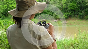a woman in a safari hat tlooking through binoculars of antelopes drinking water
