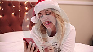 A woman in a santa claus hat lies on the bed and looks at a mobile phone, prints messages and congratulations. Christmas