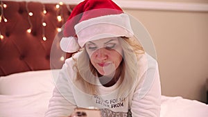 A woman in a santa claus hat lies on the bed and looks at a mobile phone, prints messages and congratulations. Christmas