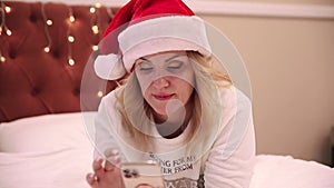 A woman in a santa claus hat lies on the bed and looks at a mobile phone, prints messages and congratulations. Christmas