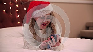 A woman in a santa claus hat lies on the bed and looks at a mobile phone, prints messages and congratulations. Christmas