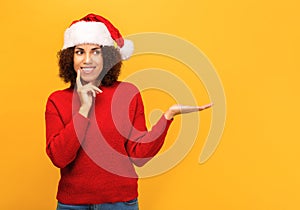 woman in a santa claus hat stands on orange background with an open palm pointing at empty space. Christmas, xmas