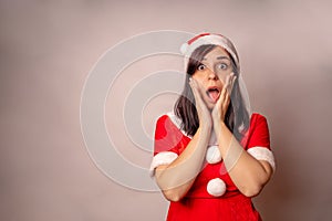 Woman in Santa Claus suit opens mouth and clutches her cheeks in surprise on grey background. Close up of female in