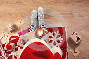 Woman in santa hat drinking cappuccino coffee and sitting on the wooden floor. Close-up of female legs in warm socks with a deer w