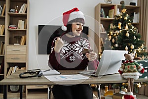 Woman in Santa hat raising fist while looking at laptop with credit card in hand