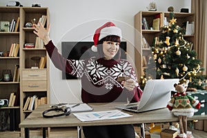 Woman in Santa hat raising palm hand while looking at laptop with credit card.