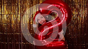 Woman in santa hat with red figure two on background of shimmering golden tinsel. Flirtatious female looks out of