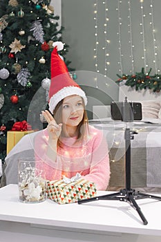 Woman in santa hat showing greeting or victory sign by video call, modern phone technology at New Year or Christmas