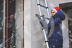 Woman with santa hat standing on ladder and decorating house exterior with outdoor christmas string lights photo