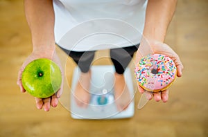 Woman on scale measuring weight holding apple and donuts choosing between healthy or unhealthy food