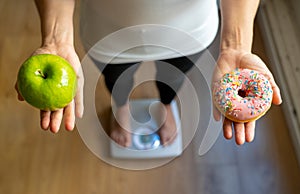 Woman on scale measuring weight holding apple and donuts choosing between healthy or unhealthy food