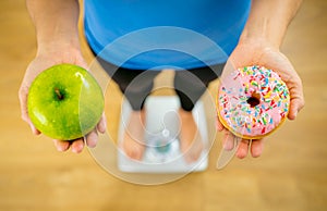 Woman on scale measuring weight holding apple and donuts choosing between healthy or unhealthy food