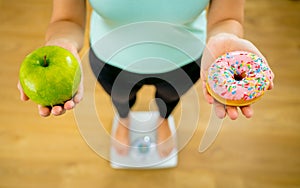 Woman on scale measuring weight holding apple and donuts choosing between healthy or unhealthy food