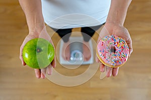 Woman on scale measuring weight holding apple and donuts choosing between healthy or unhealthy food