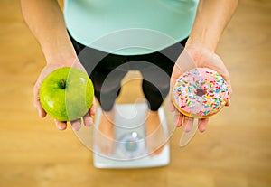 Woman on scale measuring weight holding apple and donuts choosing between healthy or unhealthy food