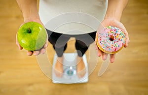 Woman on scale measuring weight holding apple and donuts choosing between healthy or unhealthy food