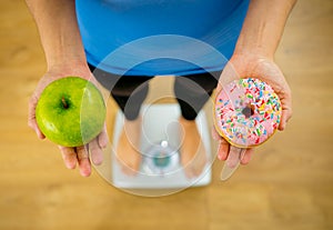 Woman on scale measuring weight holding apple and donuts choosing between healthy or unhealthy food