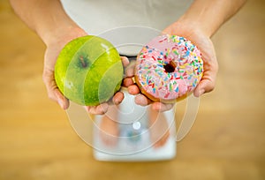 Woman on scale measuring weight holding apple and donuts choosing between healthy or unhealthy food