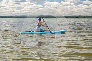 A woman in a scarf on her head and in a pareo on a SUP board with an oar swims in the lake on a sunny day.